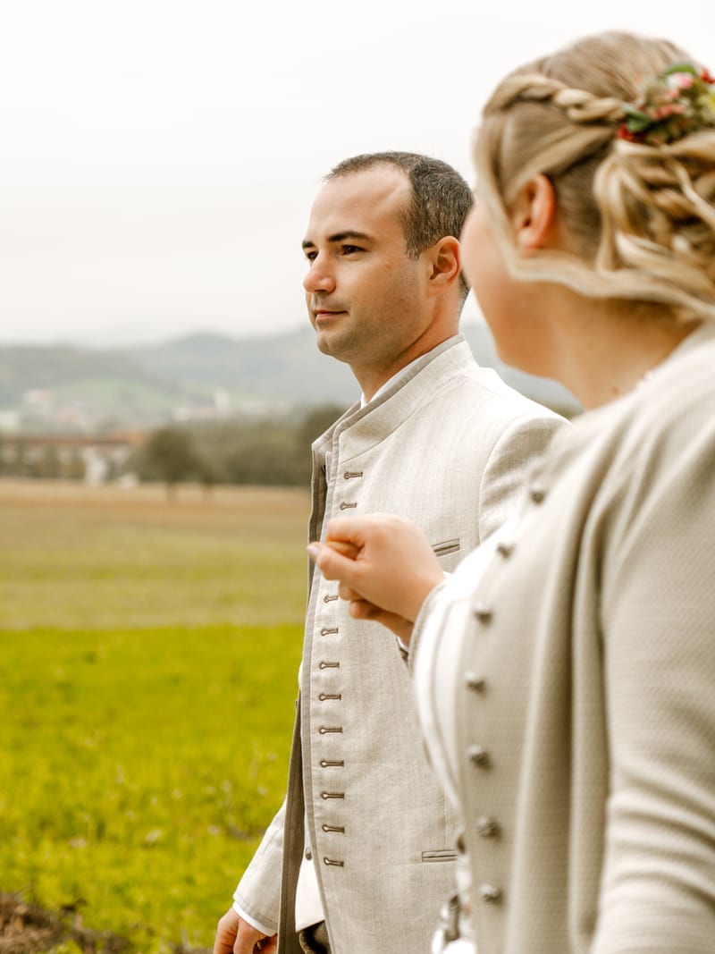 Brautpaar in Tracht auf einer Wiese, die Braut richtet dem Bräutigam die Jacke